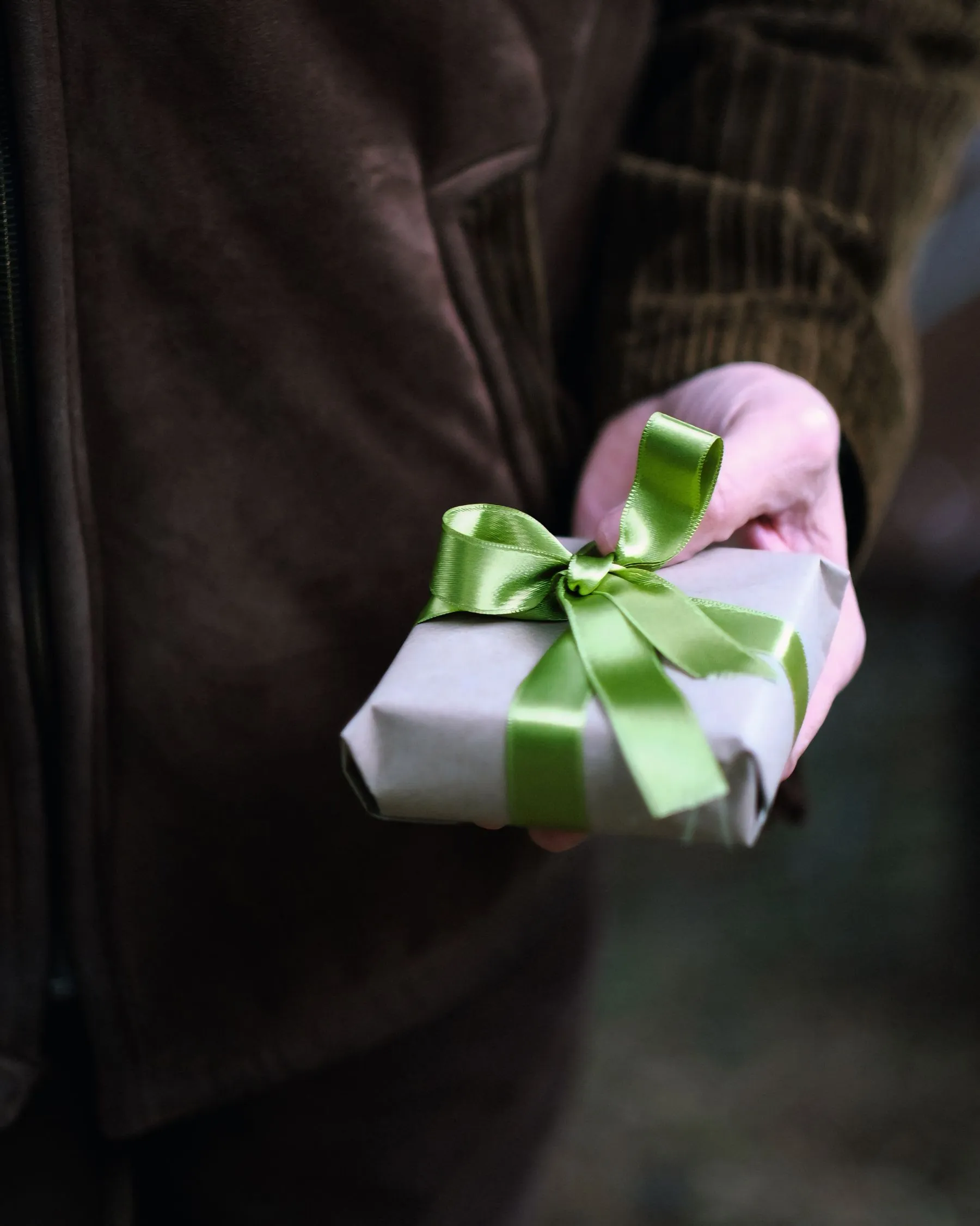 Person in a dark coat holding out a small wrapped gift with a green bow