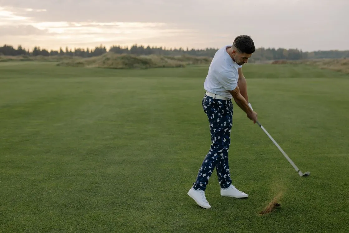 Man in a white shirt finishing an iron shot on a links fairway at dusk