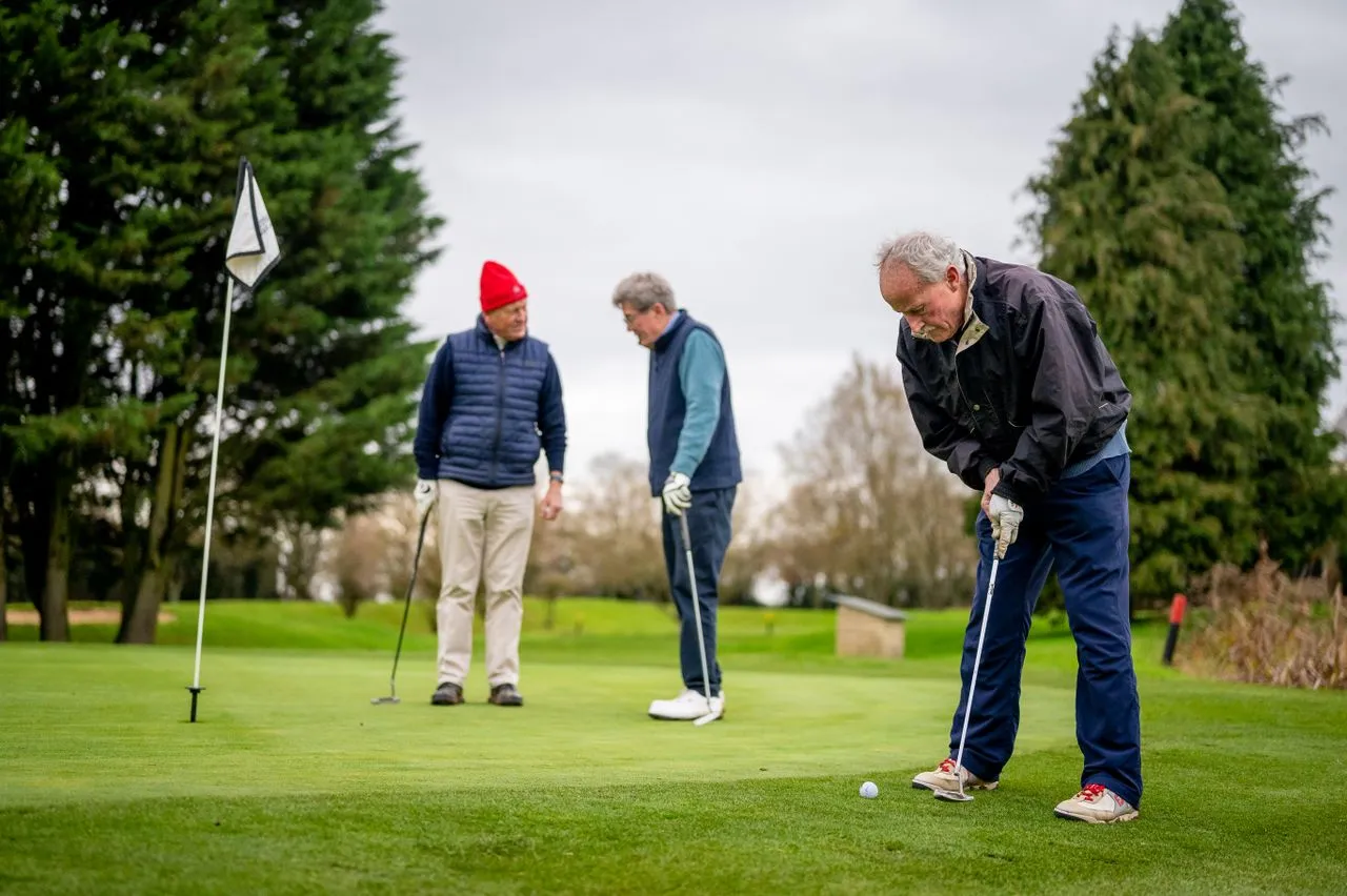 Three older friends on a putting green in winter jackets, one lining up a putt beside the flag