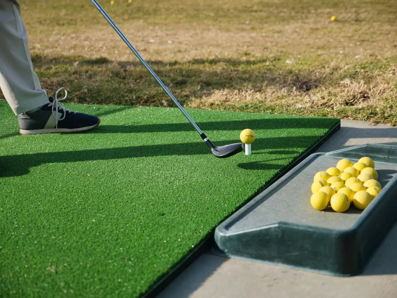 Iron behind a teed ball on an artificial hitting mat, a tray of yellow practice balls beside it